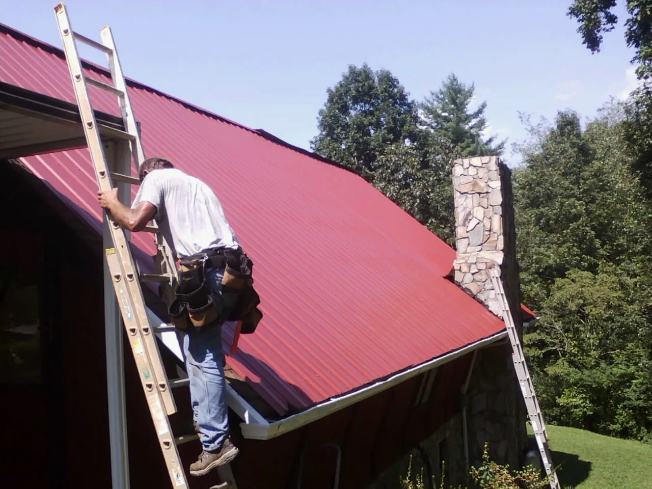 Roofer on a ladder beside a red metal roof and stone chimney — Soffit & Fascia Repair by Roofing Brothers in Osseo